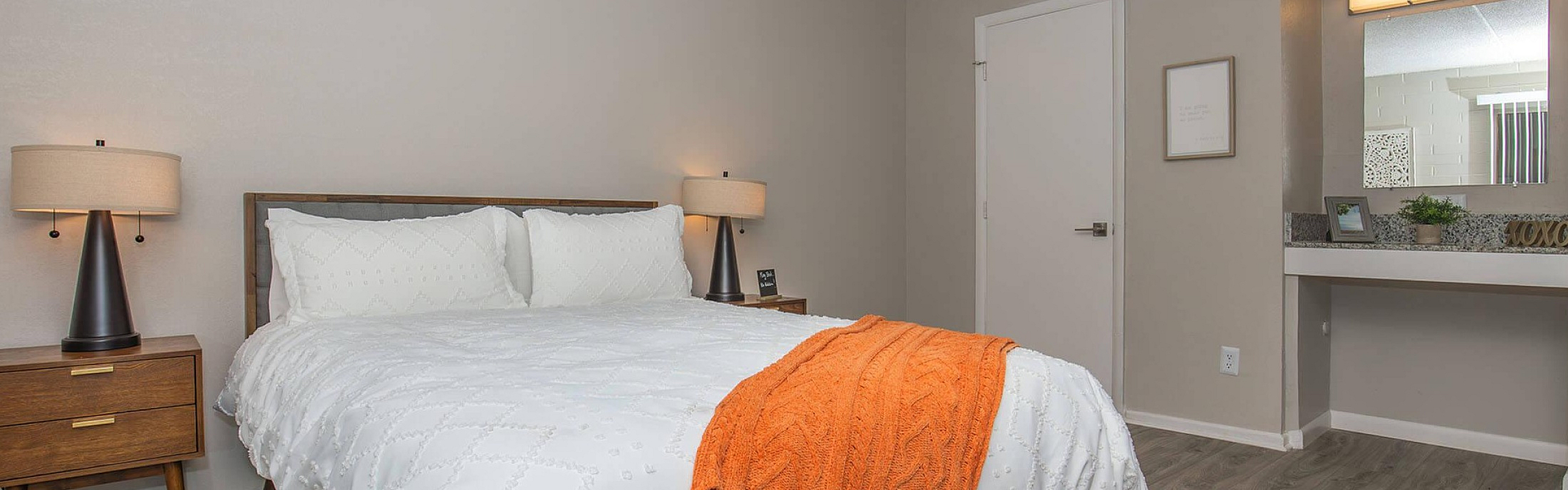 Inviting bedroom with modern decor, featuring a plush bed adorned with a vibrant orange throw, elegant bedside tables with chic lamps, and stylish wood flooring.