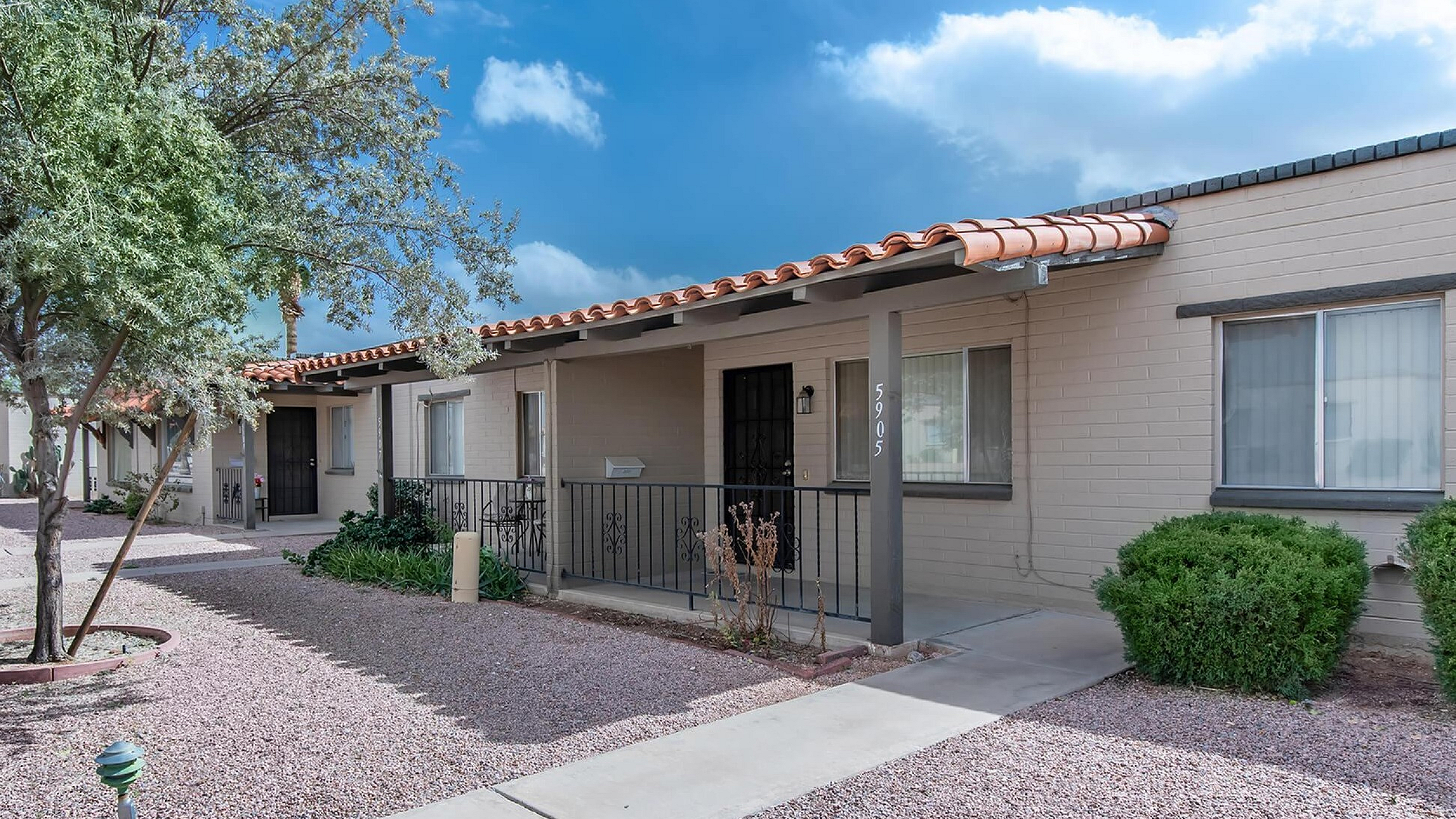 Charming single-level apartment with terracotta-tiled roof, set amidst desert landscaping and shaded by lush trees.