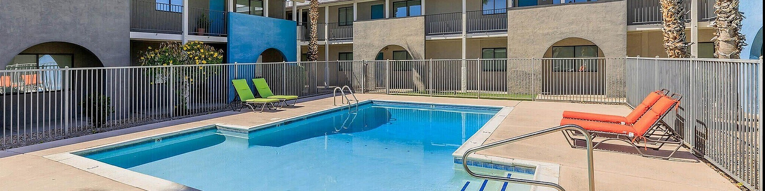 Inviting apartment complex featuring a sun-kissed swimming pool, surrounded by chic lounge chairs and vibrant palm trees, perfect for relaxation in a modern setting.