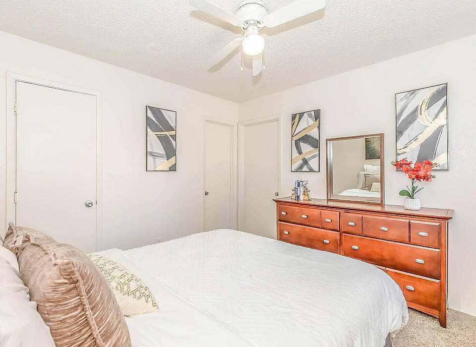 Bright and inviting bedroom with modern art, a sleek ceiling fan, and a stylish wooden dresser topped with vibrant decor.