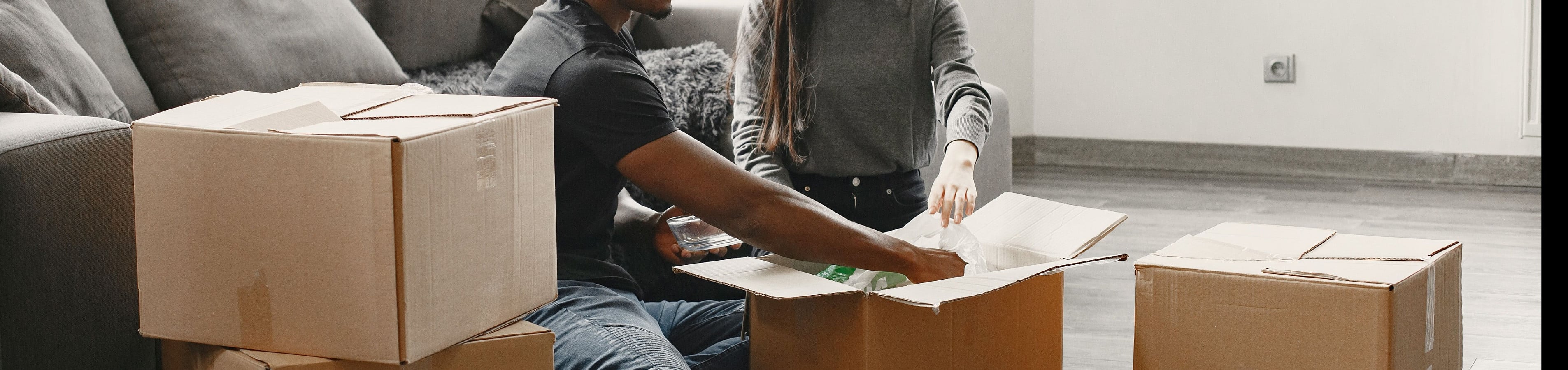Happy couple unpacking boxes in a bright, modern living room with cozy gray furniture and sleek hardwood floors, showcasing the welcoming atmosphere of the apartment.