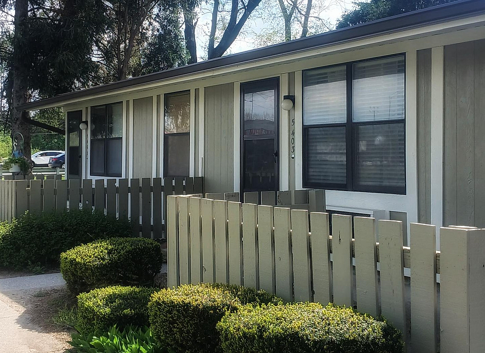 Charming single-story apartment with a modern exterior, surrounded by lush greenery and a quaint wooden fence, offering serene living.