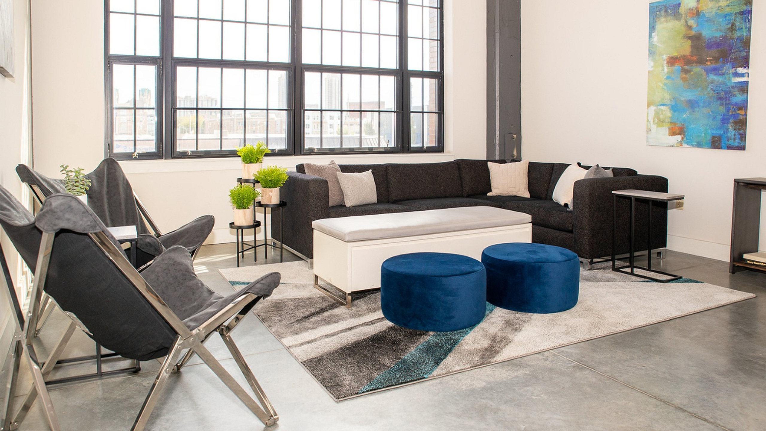 Bright and modern living space featuring a sleek black sectional, plush white ottoman, and vibrant blue stools beneath large industrial-style windows.