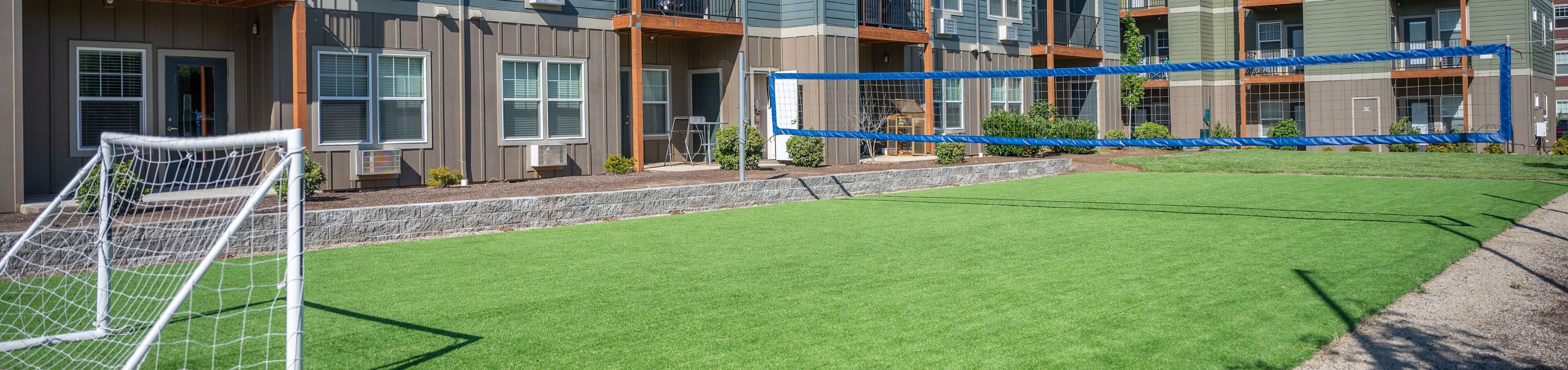 Modern apartment complex with a vibrant outdoor sports area featuring a soccer goal and volleyball net on lush green turf.