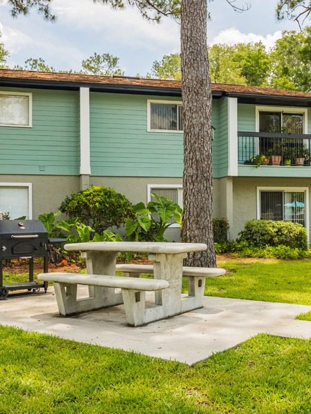 community picnic table next to grill and grass area with apartment building in the background