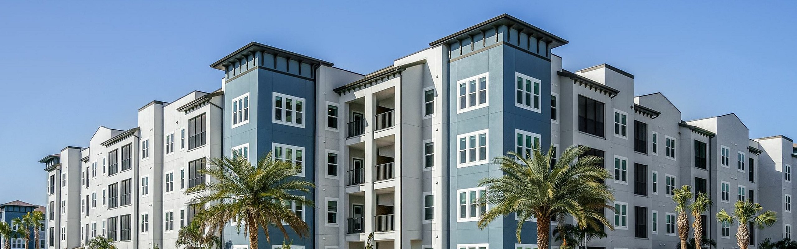 Modern apartment complex with vibrant blue and white facade, surrounded by palm trees and lush landscaping under a clear blue sky.