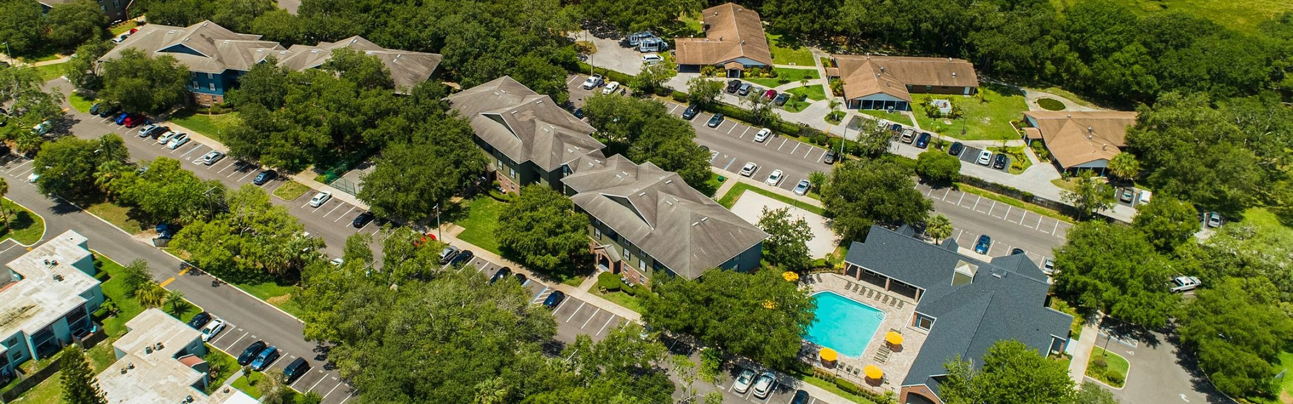 Aerial view of a vibrant apartment complex surrounded by lush greenery, featuring multiple residential buildings, spacious parking areas, and a sparkling blue swimming pool with sun umbrellas.