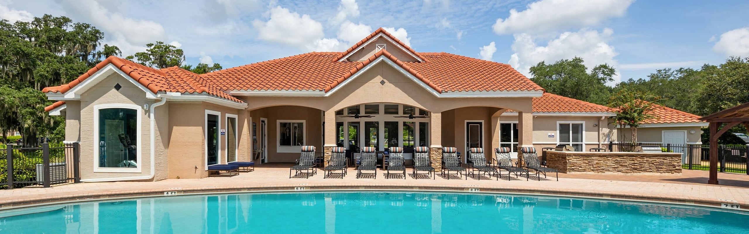 Elegant residential complex featuring a stunning swimming pool with a serene setting under a bright blue sky, complemented by stylish sun loungers and a charming building with terracotta roofs.