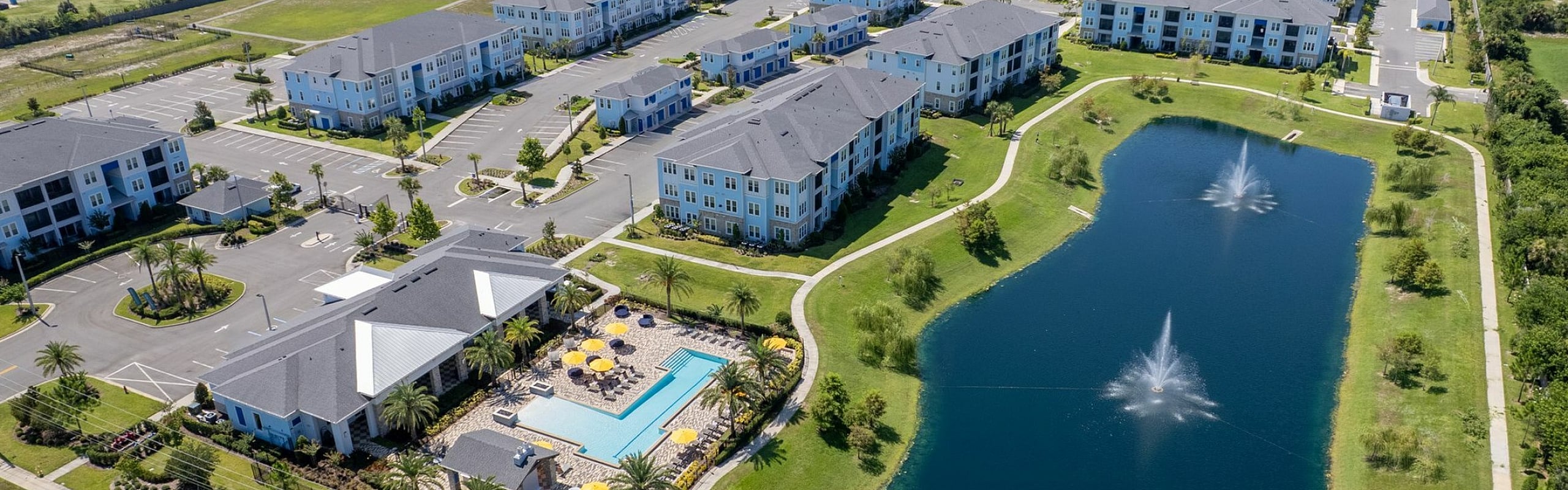 Aerial view of a vibrant apartment complex featuring modern buildings, a pristine pool area with yellow umbrellas, and a shimmering lake with fountains, surrounded by lush greenery.