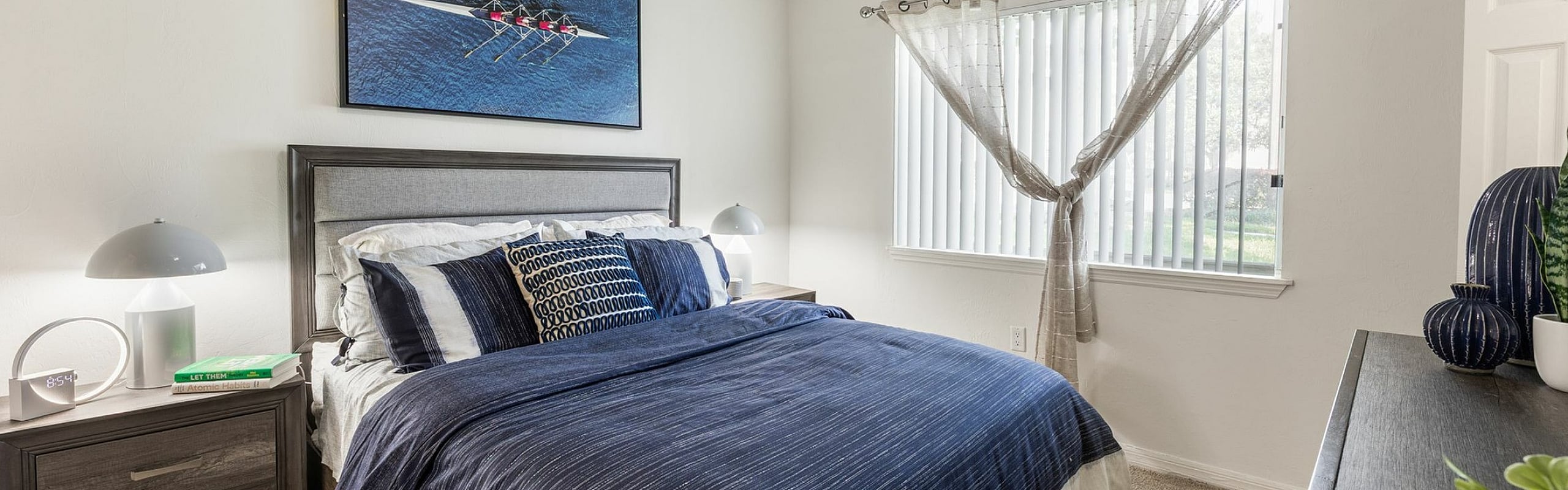 Bright and inviting bedroom with modern decor, featuring a plush bed with navy accents, stylish artwork, and abundant natural light through large windows.