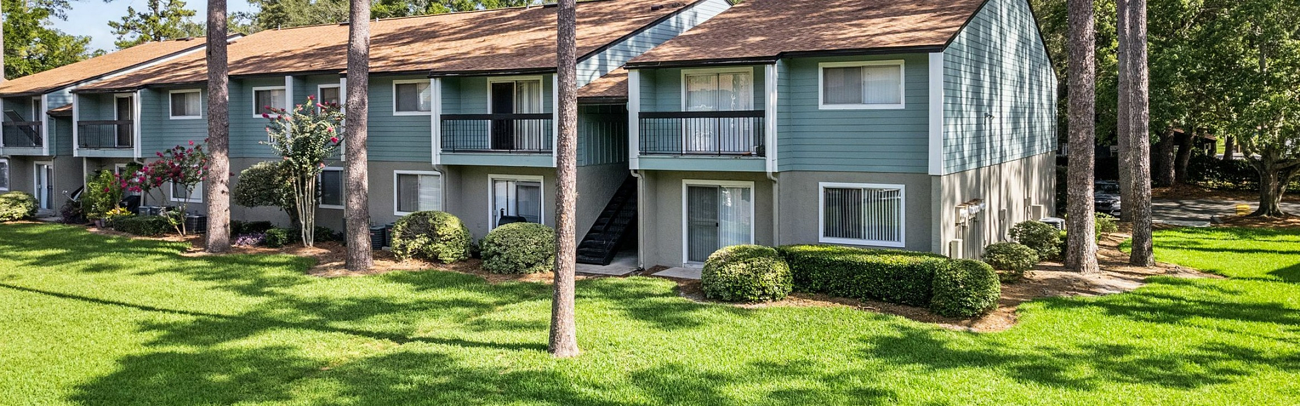 Charming apartment complex surrounded by lush greenery and tall pine trees, featuring light blue two-story buildings with private balconies and a serene garden setting.