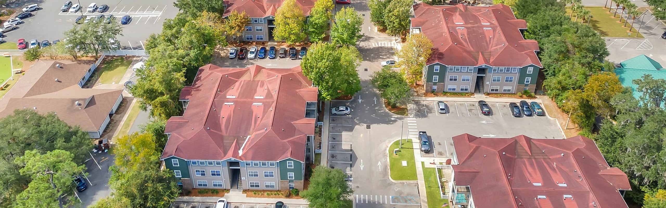 Vibrant aerial view of a well-planned residential community surrounded by lush green trees, featuring modern buildings with red roofs, ample parking, and nearby amenities.