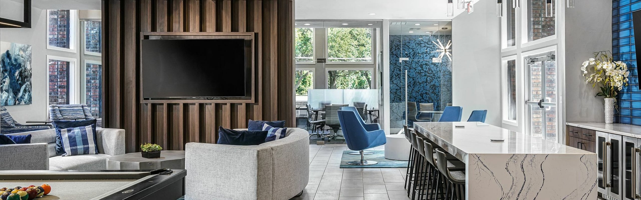 Modern communal entertainment space featuring a sleek pool table, stylish seating area with gray sofas, large TV, and a chic marble kitchen island with seating. Natural light floods the room through expansive windows, highlighting the conte