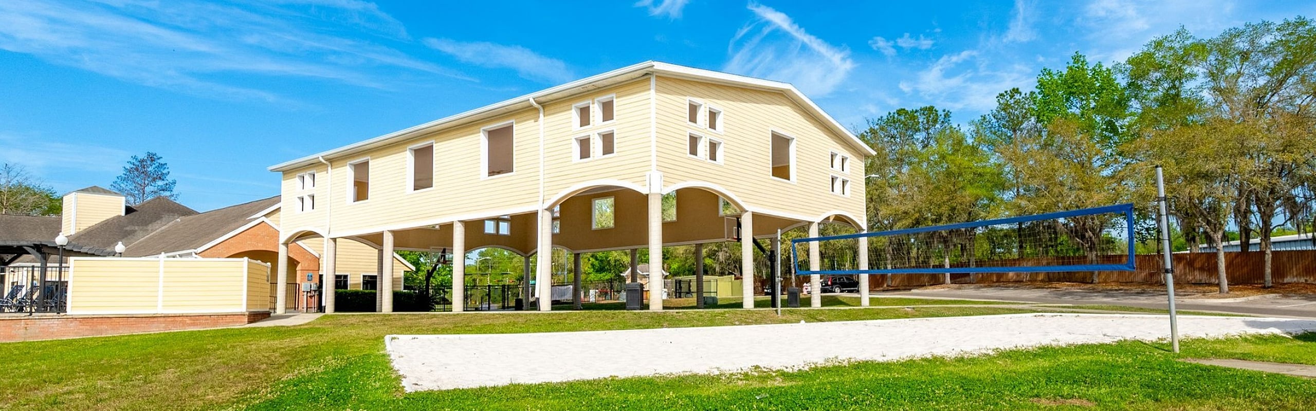 Bright residential building with elevated design, surrounded by lush greenery and featuring a sand volleyball court against a clear blue sky.