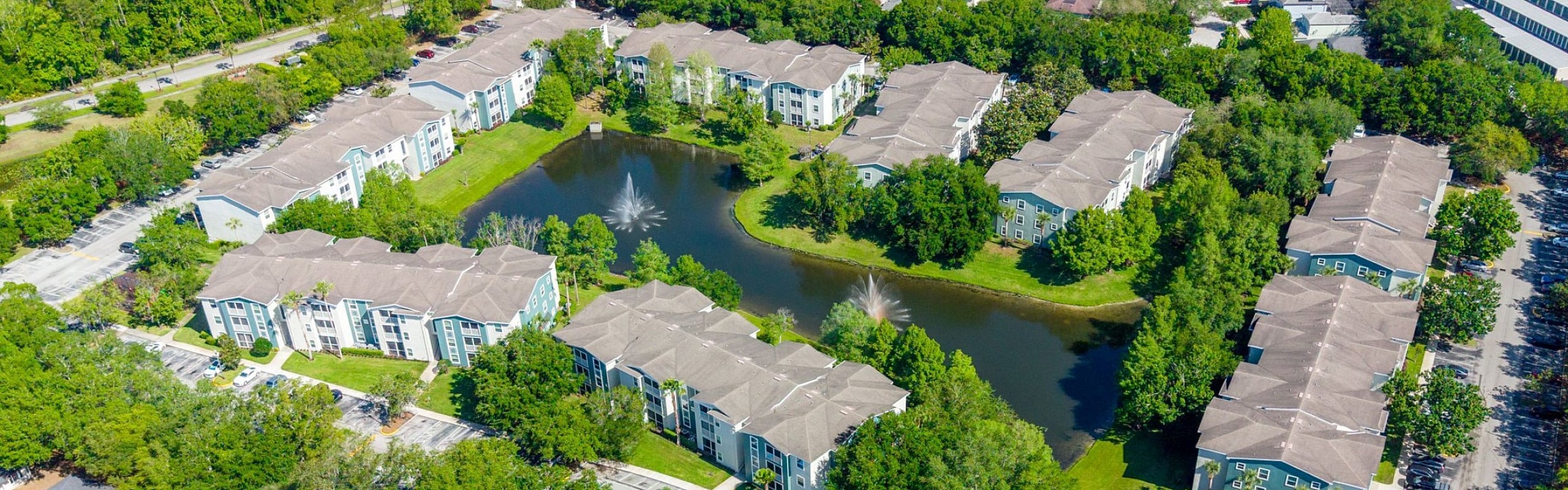 Aerial view of a vibrant apartment complex featuring lush greenery, a serene pond with fountains, community tennis courts, and a refreshing swimming pool.