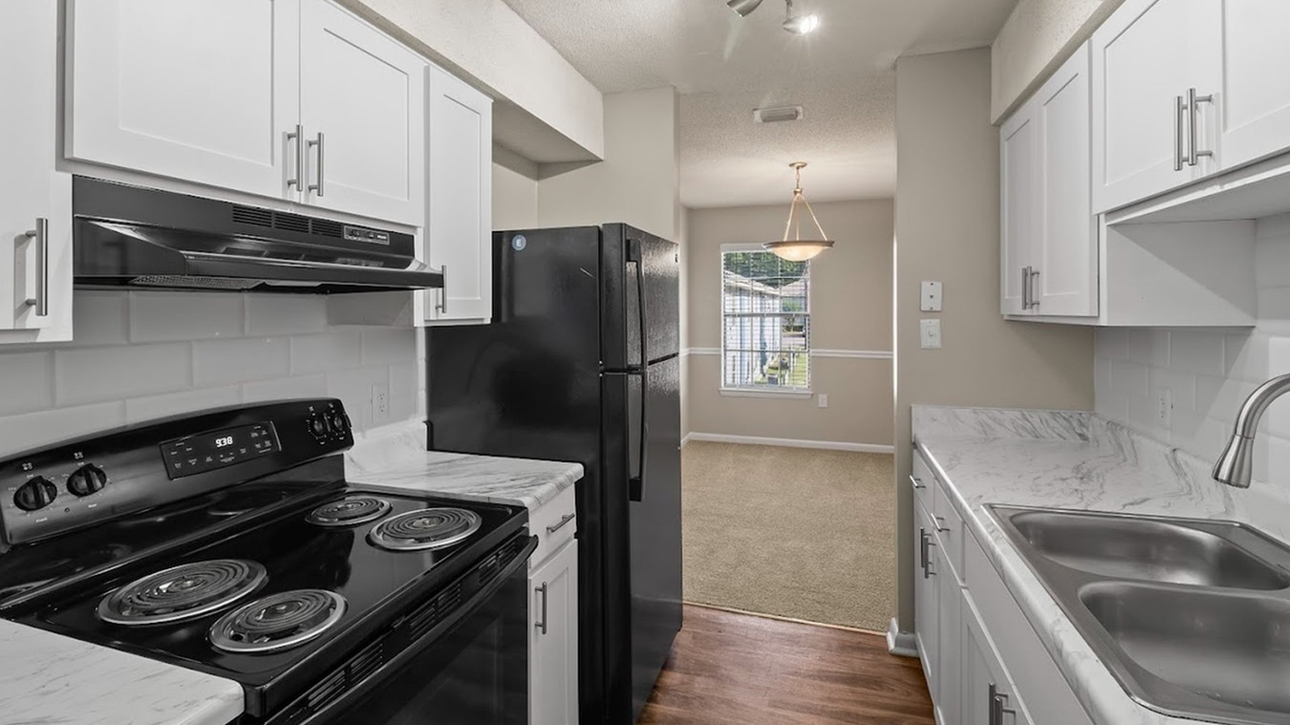 Modern kitchen with sleek white cabinets, black appliances, and hardwood flooring leading to a bright dining area.