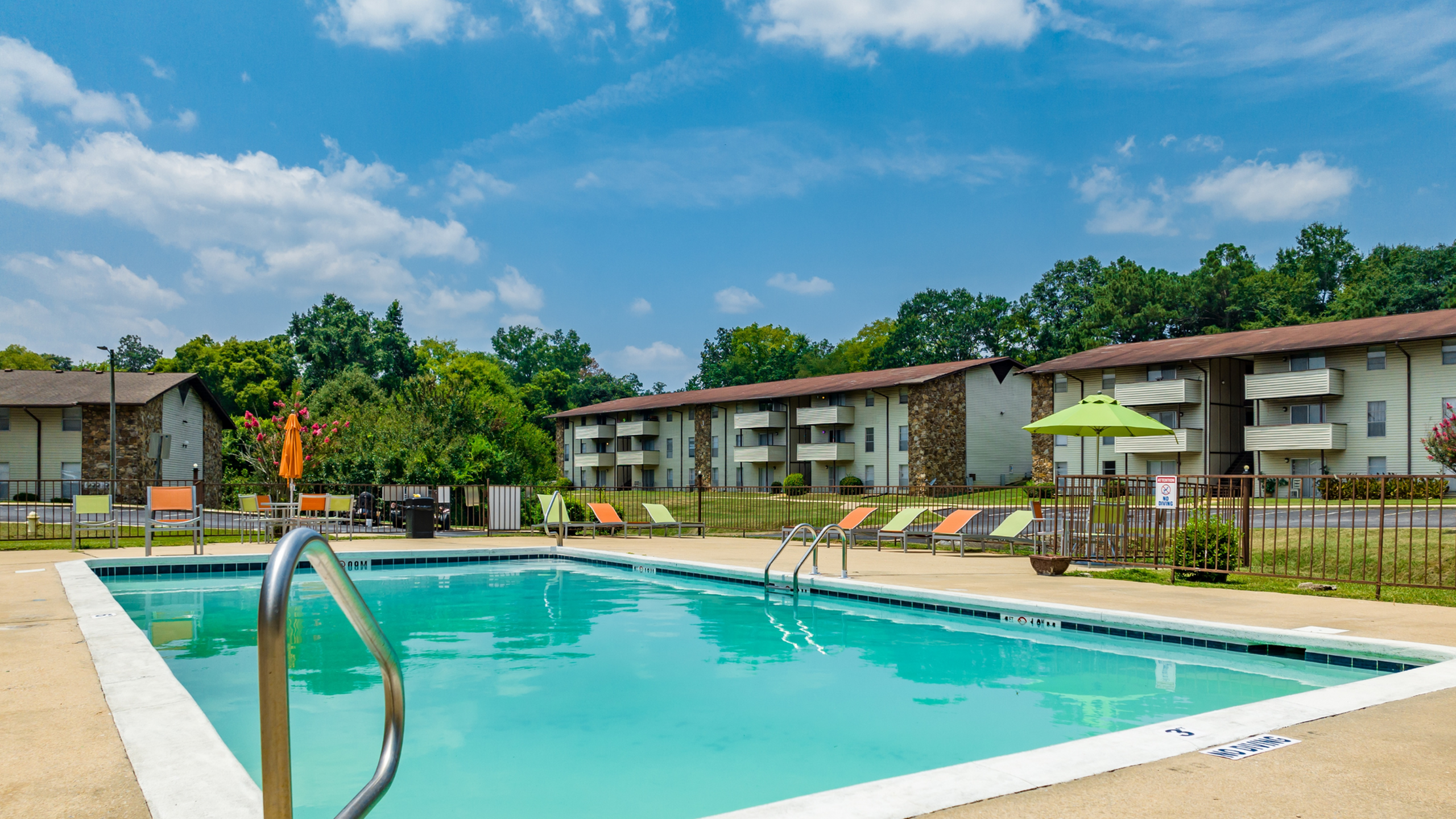 Inviting apartment complex featuring a sparkling outdoor pool, colorful lounge chairs, and stunning landscaping under a clear blue sky.