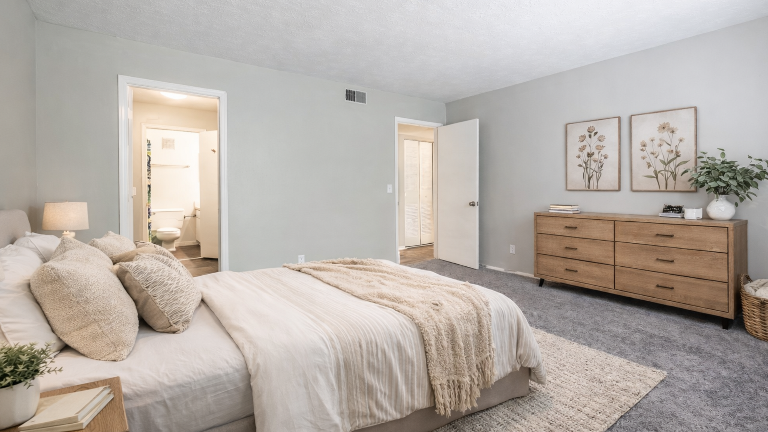 Elegant bedroom featuring a cozy queen bed with textured pillows, soft lighting, and a chic wooden dresser with floral art decor.