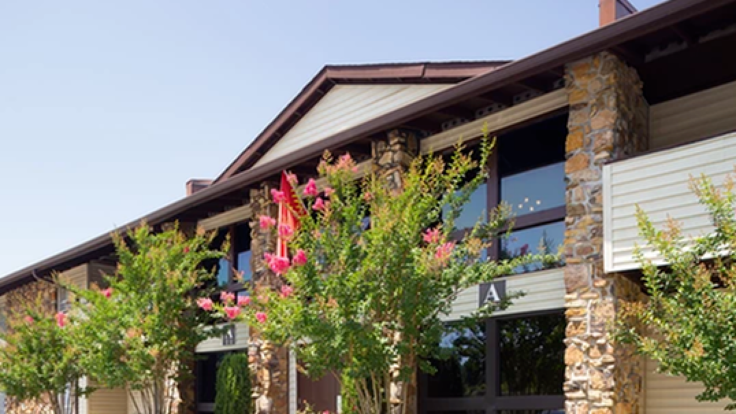 Charming apartment exterior with stone accents and lush landscaping, showcasing vibrant flowering trees under a clear blue sky.