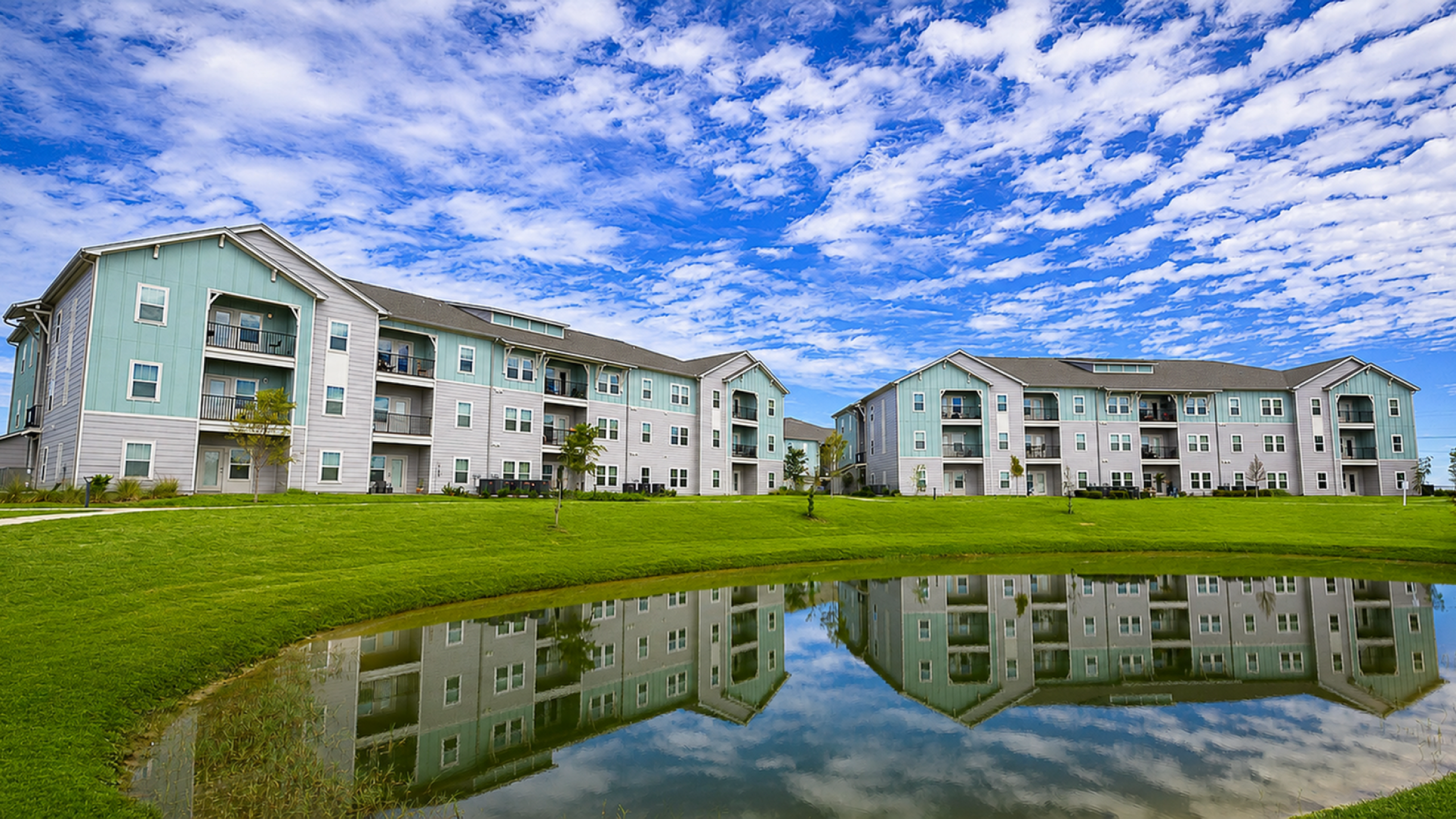 Modern apartment complex with pastel green accents, reflecting in a serene pond under a vibrant blue sky.