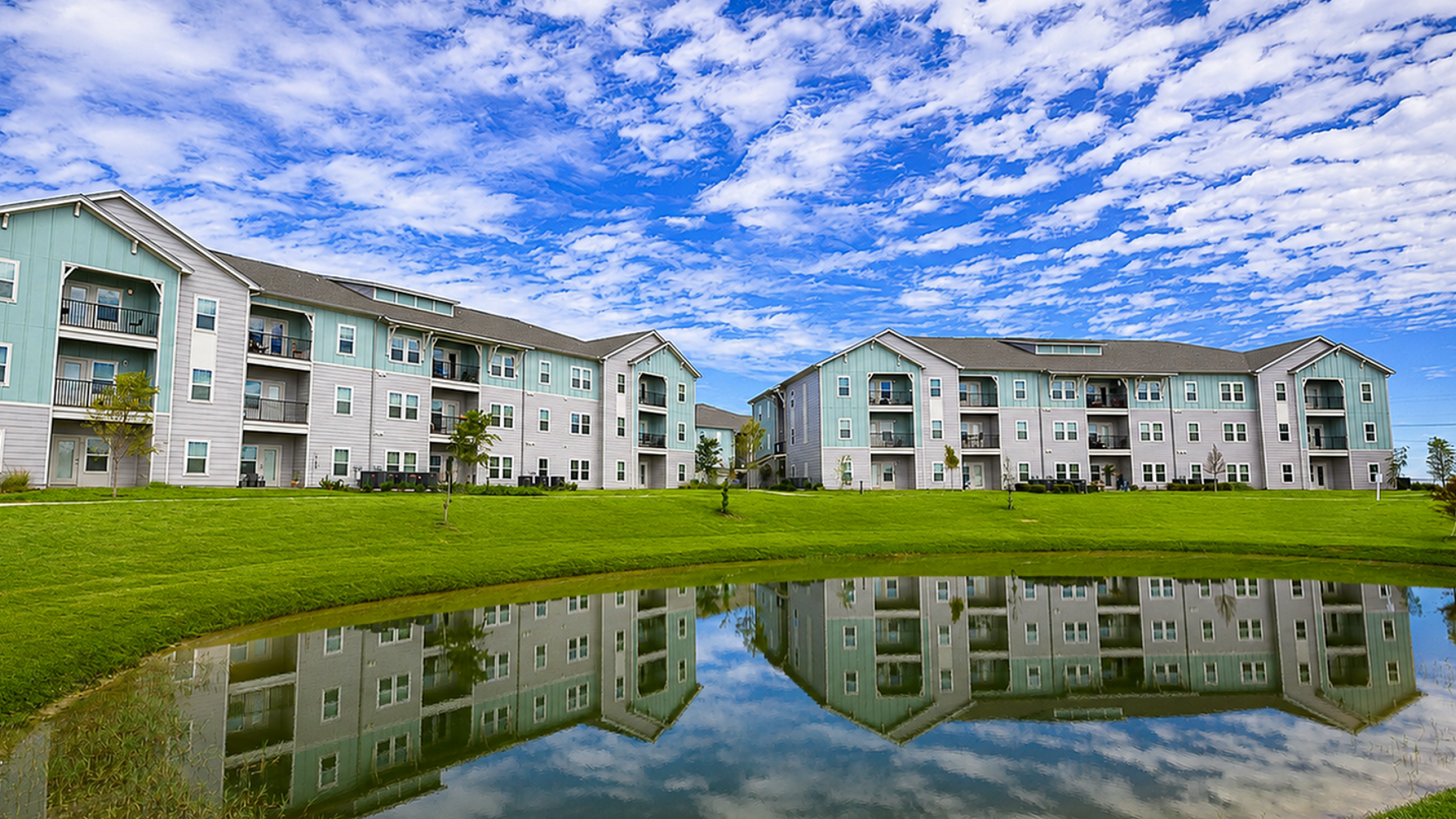Charming modern apartment complex with serene pond and vibrant green lawn, set against a beautiful blue sky with fluffy clouds.