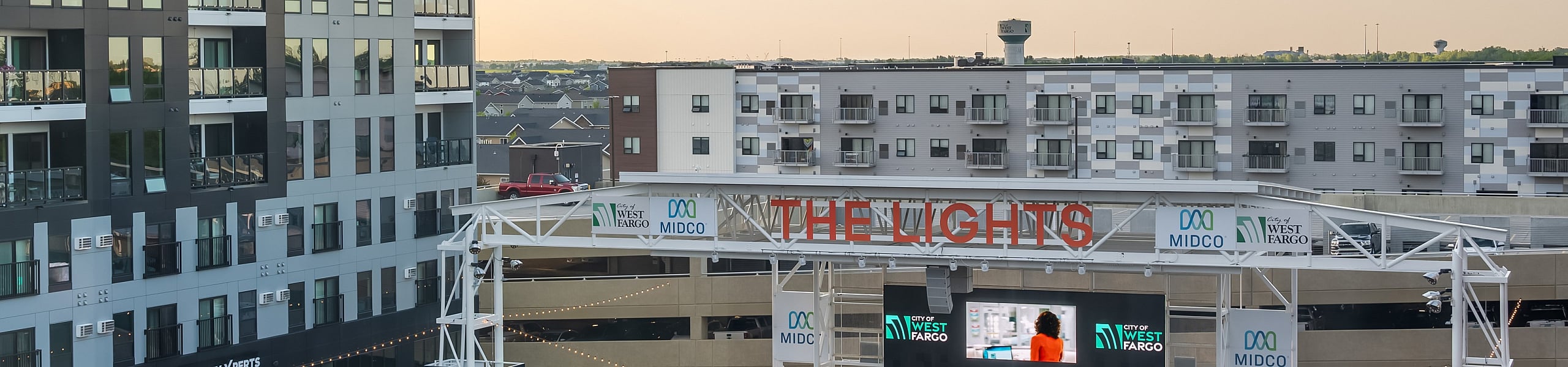 Modern urban apartment complex overlooking a vibrant outdoor entertainment venue with a spacious event area, surrounded by stylish architecture at sunset.