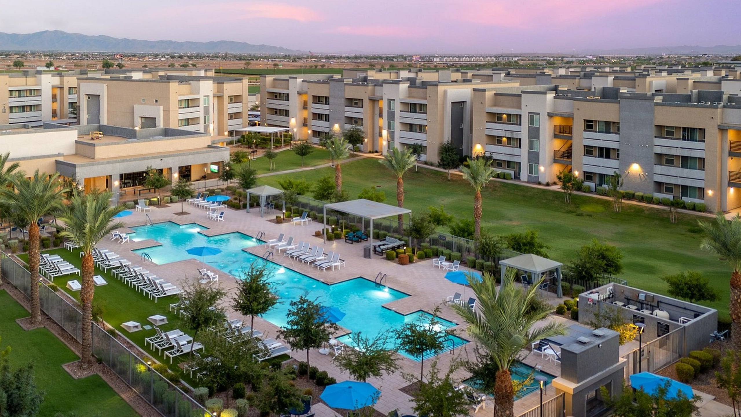 Luxurious apartment complex with stunning resort-style pool, surrounded by lush greenery and modern buildings at sunset.