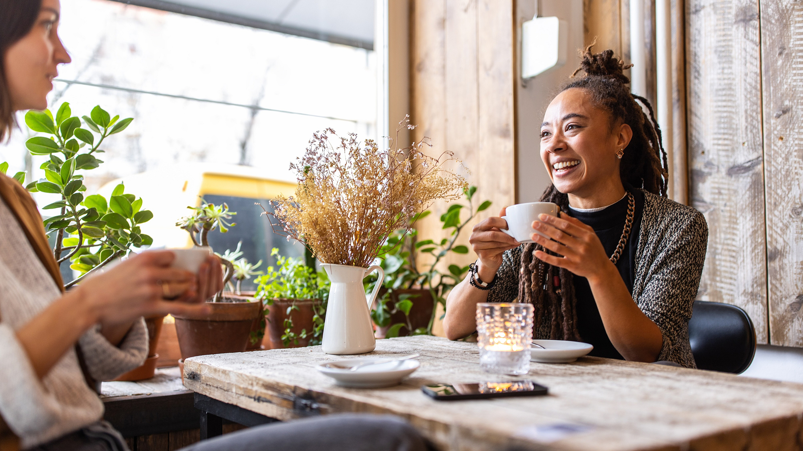 Charming cafe scene with two friends enjoying coffee amidst lush green plants and rustic decor.