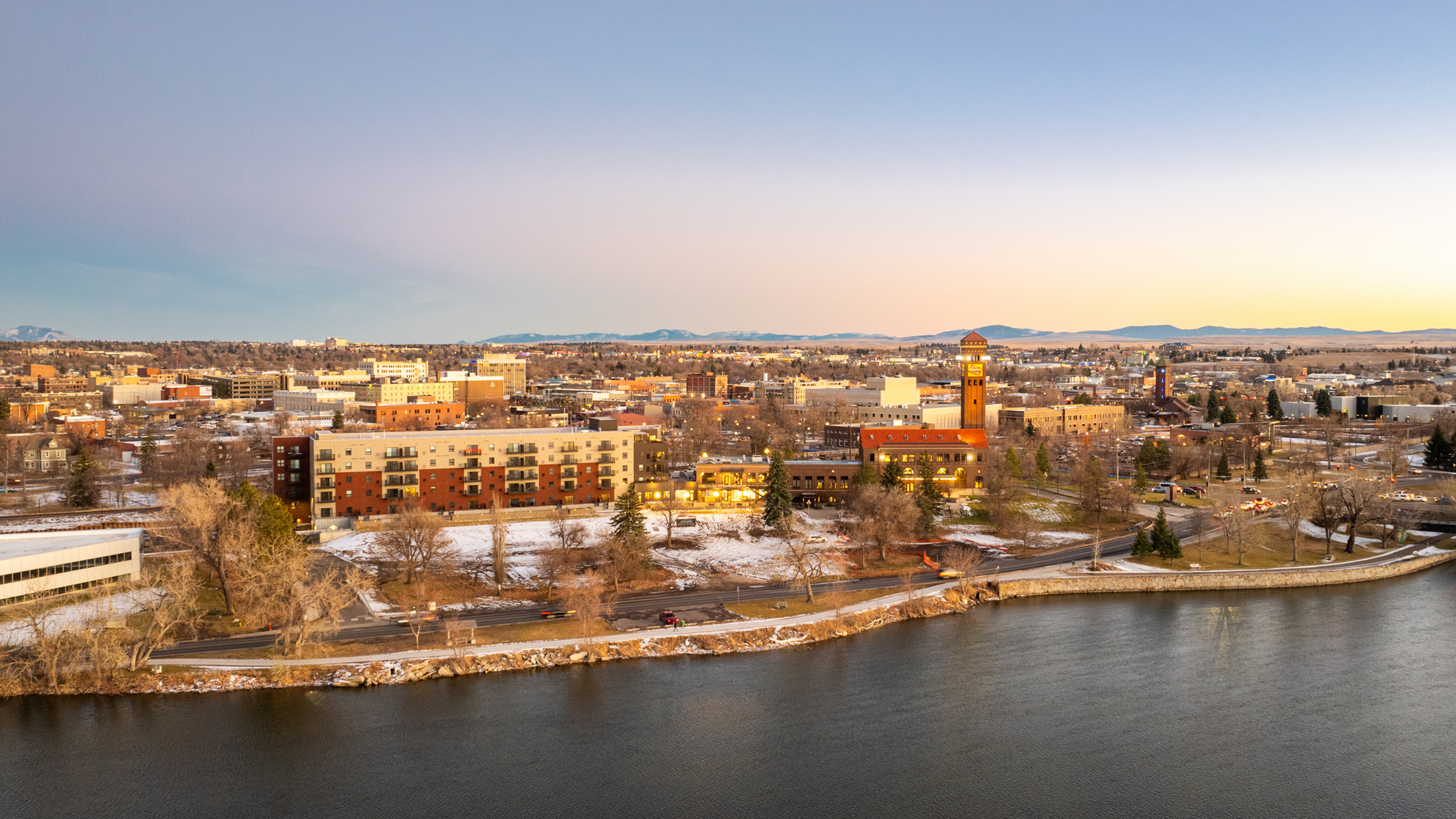 Panoramic view of a cityscape featuring modern buildings along a riverside at sunset.