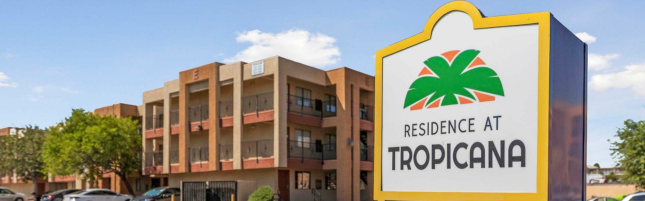Charming apartment complex with a prominent sign reading 'Residence at Tropicana', surrounded by lush greenery and parked cars.