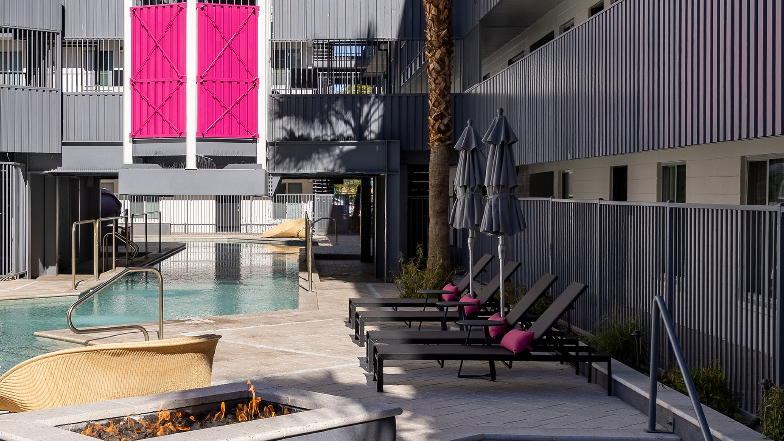 Modern apartment complex with vibrant pink accents, featuring a sleek pool area, sun loungers, and a cozy fire pit under clear blue skies.