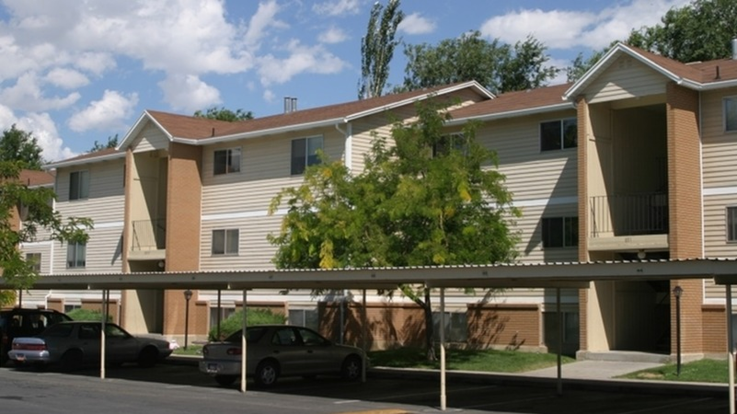 Charming apartment complex with covered parking, surrounded by lush greenery and bathed in sunlight.
