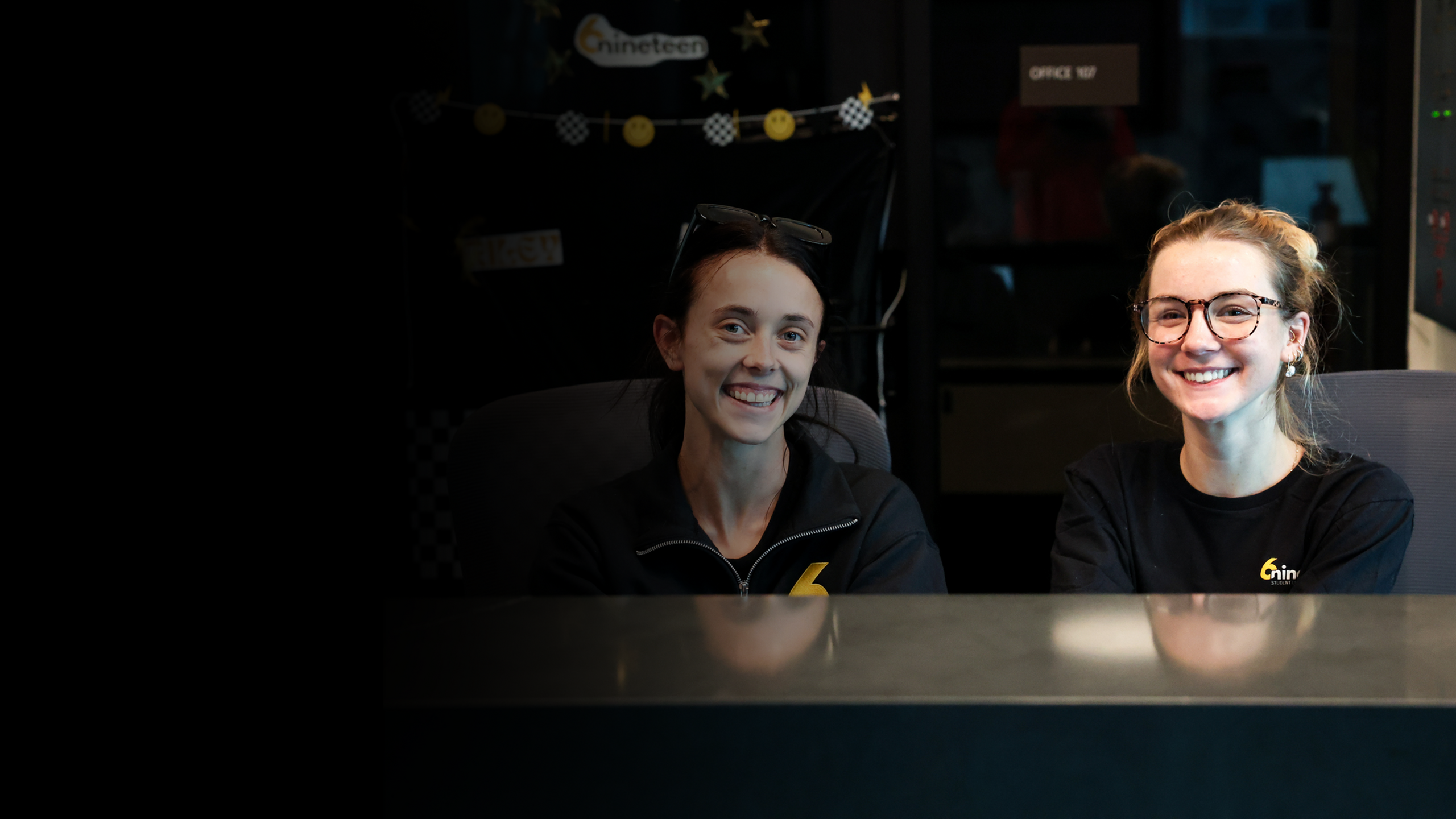 Two smiling front-desk team members sit behind the leasing counter in a modern student-housing office, creating a welcoming first impression for prospects. Their branded apparel and upbeat atmosphere highlight the community&rsquo;s friendly