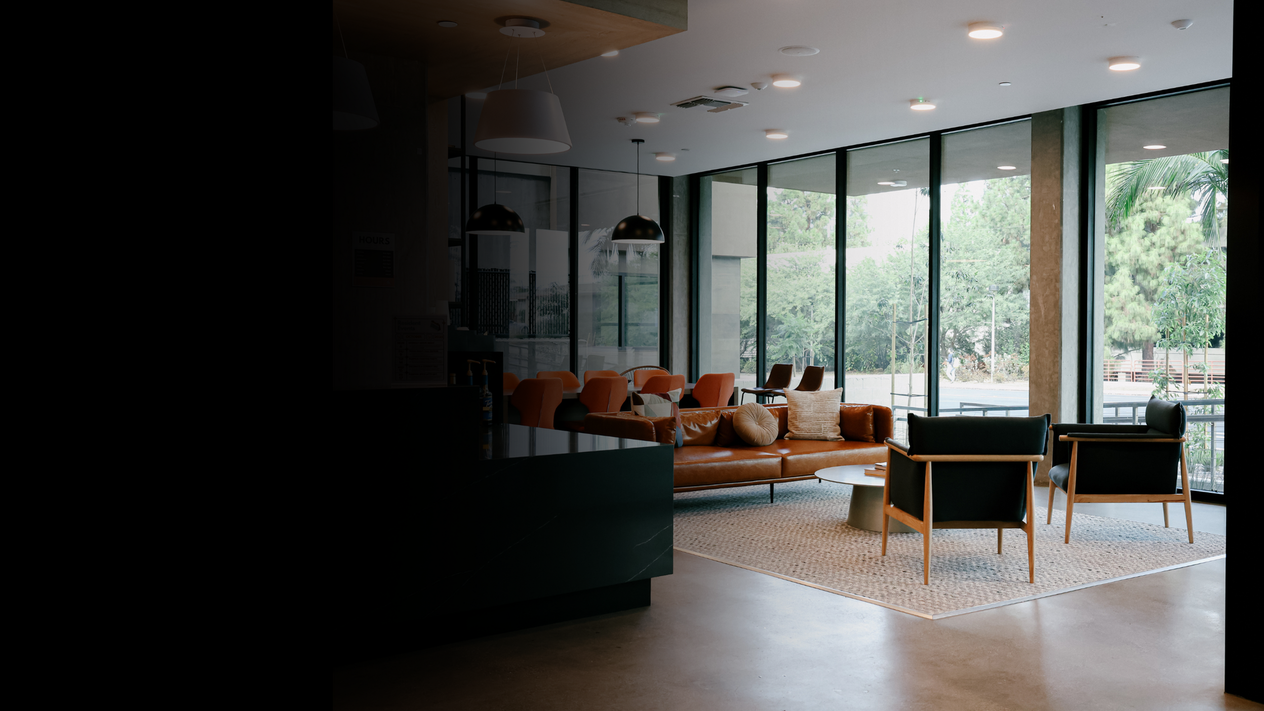 Modern apartment clubhouse lounge with a leather sofa, accent chairs, and floor-to-ceiling windows overlooking greenery, offering a stylish and comfortable community space for residents to relax or socialize.