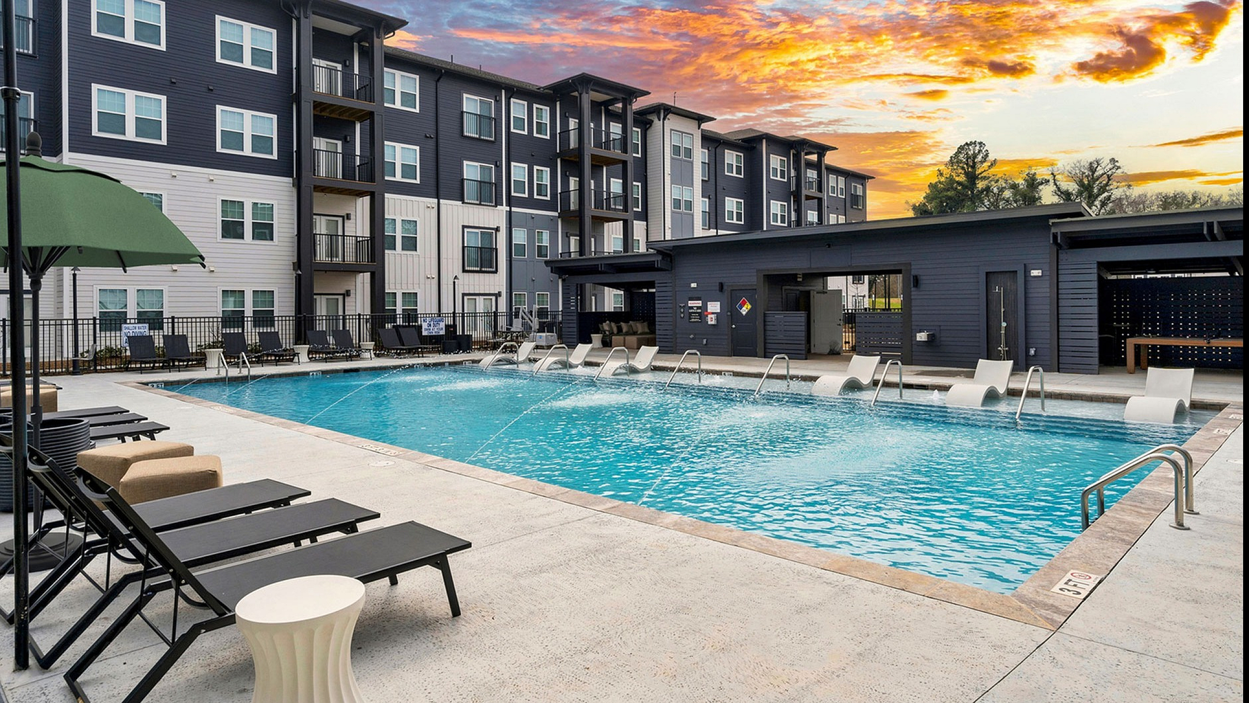 Stunning apartment complex with modern design, featuring a luxurious outdoor swimming pool and relaxing lounge chairs, set against a vibrant sunset sky.