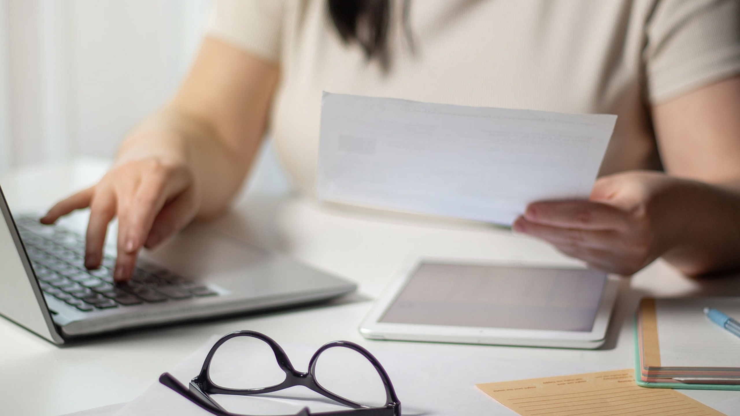 Focused workspace showing a person reviewing paperwork with a laptop, tablet, and glasses on a bright, organized desk.