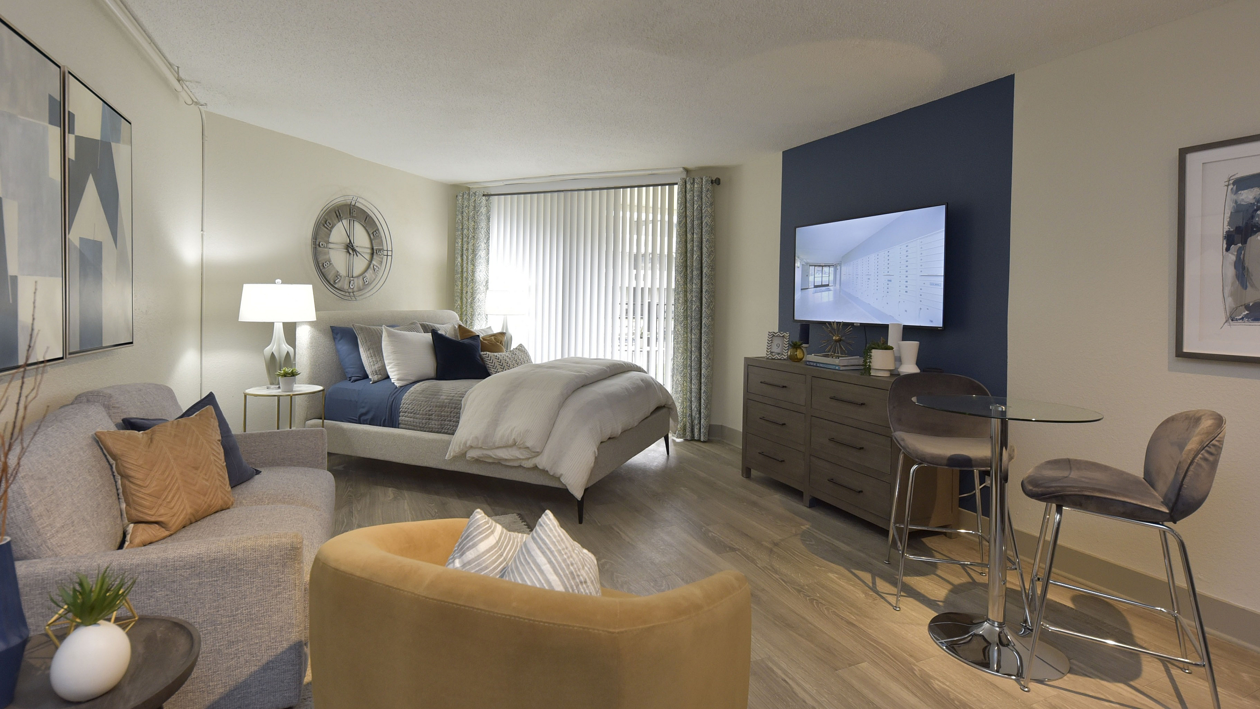 Modern studio apartment featuring a cozy sleeping area with a plush bed, stylish seating with accent chairs, a sleek dining space with a glass table, and elegant decor against a rich blue accent wall.