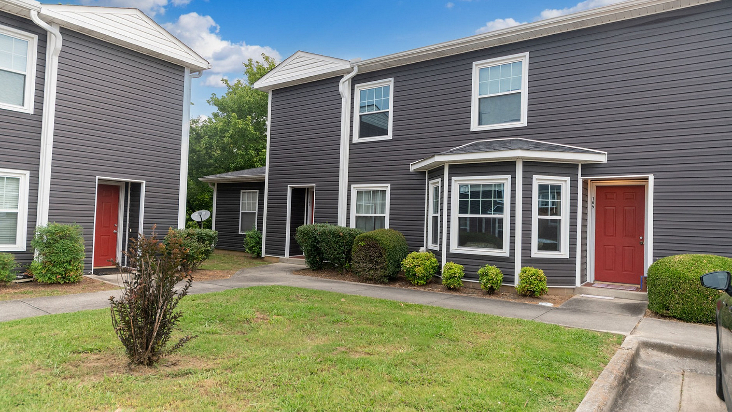 Charming townhouse with modern dark siding and vibrant red doors surrounded by lush greenery and clear blue skies, offering a welcoming neighborhood ambiance.