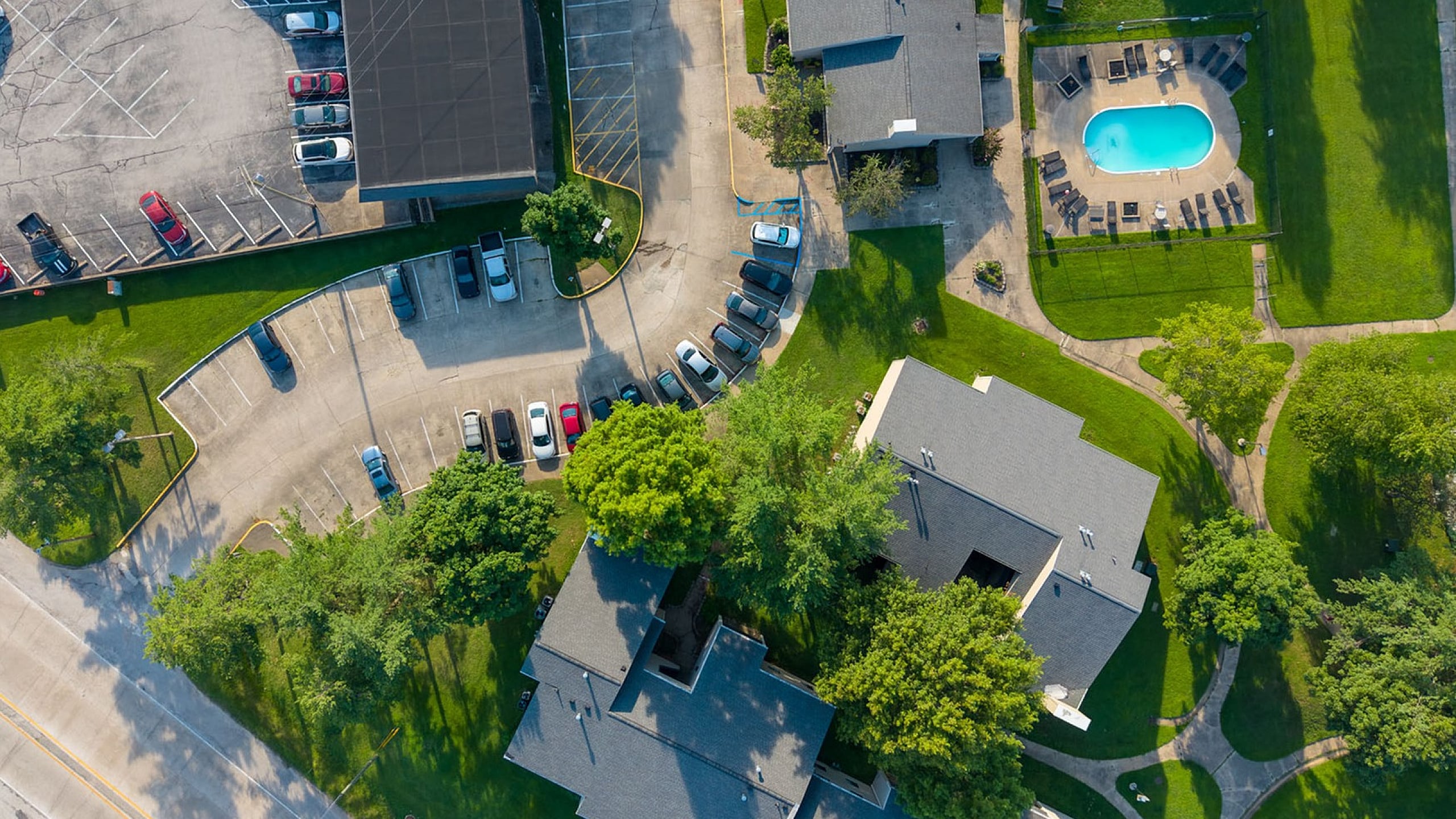 Aerial view of a vibrant apartment complex with lush green lawns, ample parking, and a sparkling blue swimming pool.