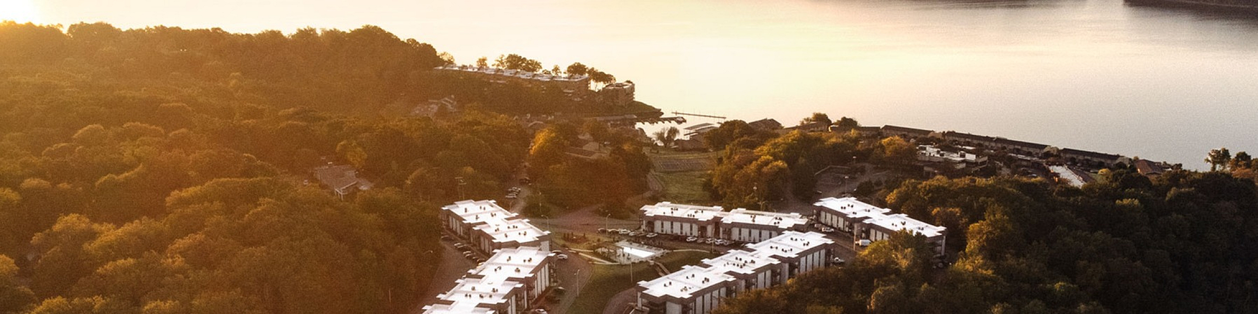 Breathtaking aerial view of modern residential community nestled amidst lush greenery, with a serene lake backdrop at sunrise.