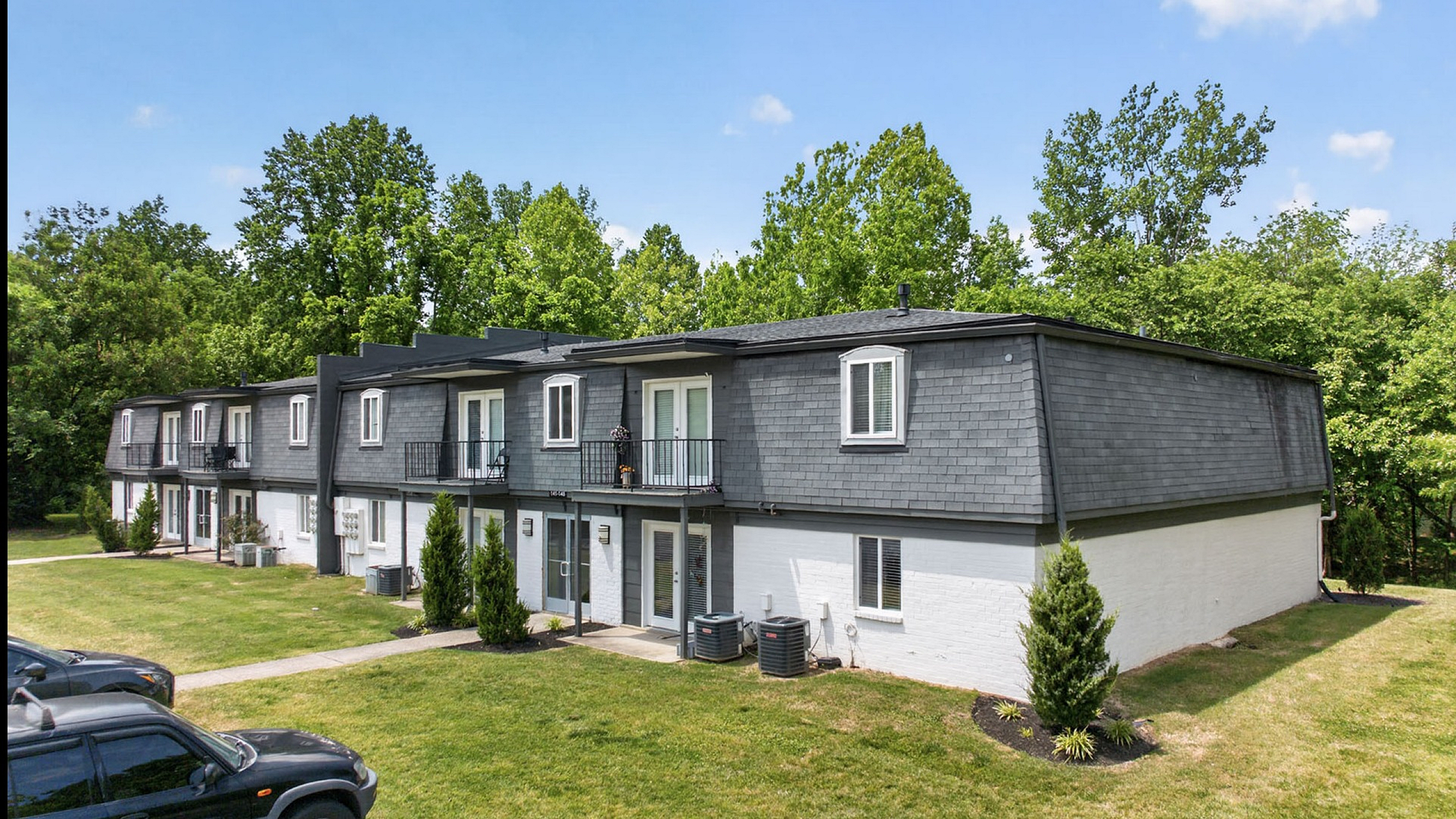 Charming townhouse complex with modern gray exteriors, lush green lawns, and inviting balconies against a backdrop of vibrant trees.