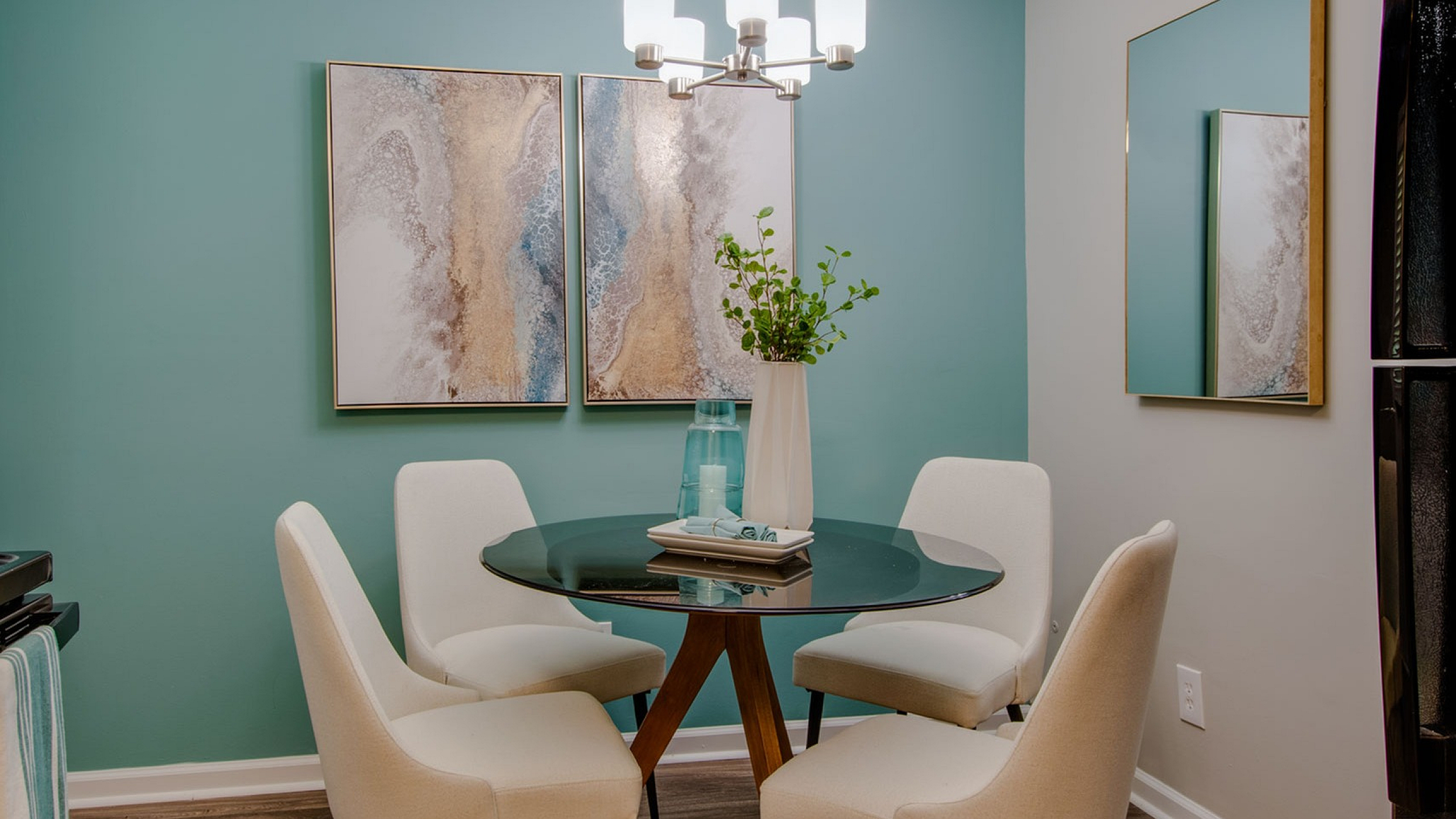 Chic dining nook featuring a round glass table, four elegant chairs, and abstract wall art against a soothing teal accent wall.