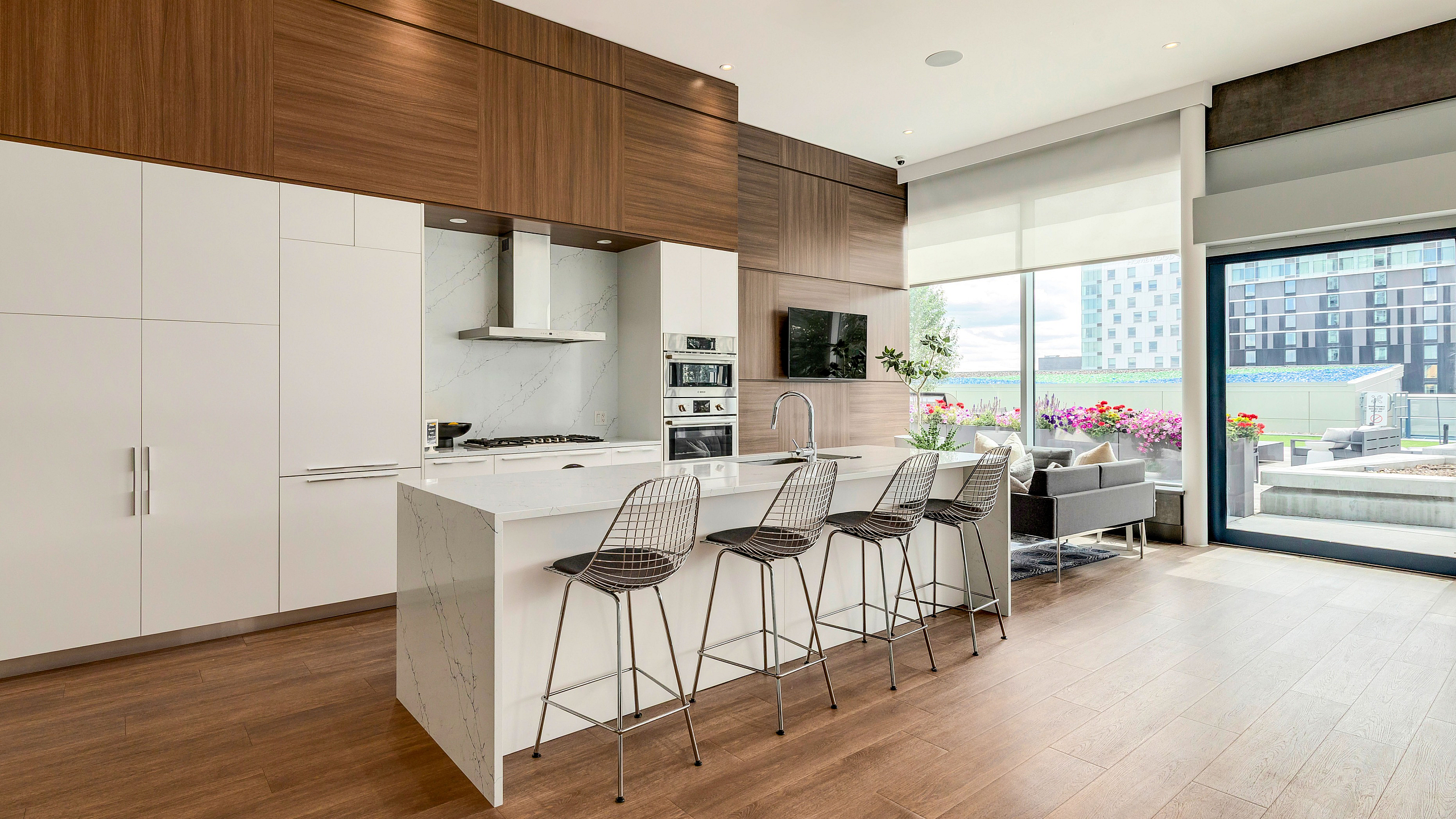 Modern kitchen with sleek white cabinetry, marble countertops, and stainless steel appliances, featuring an inviting breakfast bar with chic wire bar stools, and a view of a sunlit living area with vibrant outdoor greenery.