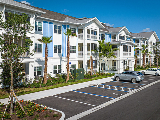 Charming modern apartment building with bright, fresh facade, surrounded by lush landscaping and ample parking, under a clear blue sky.