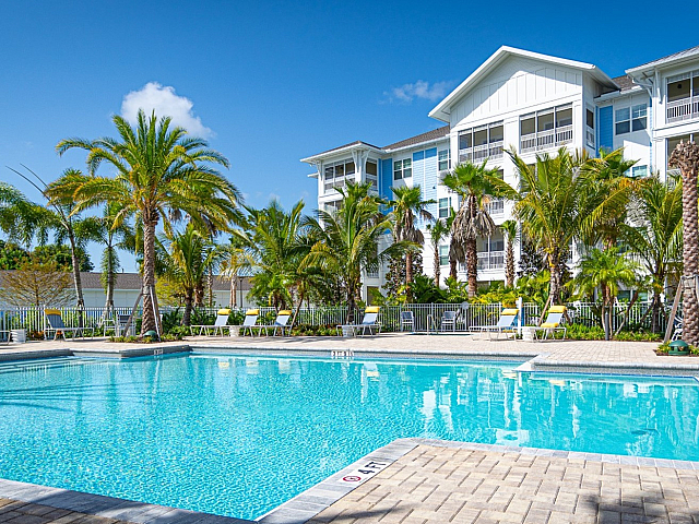 Luxurious apartment complex with sparkling pool, surrounded by lush palm trees and bright blue skies.