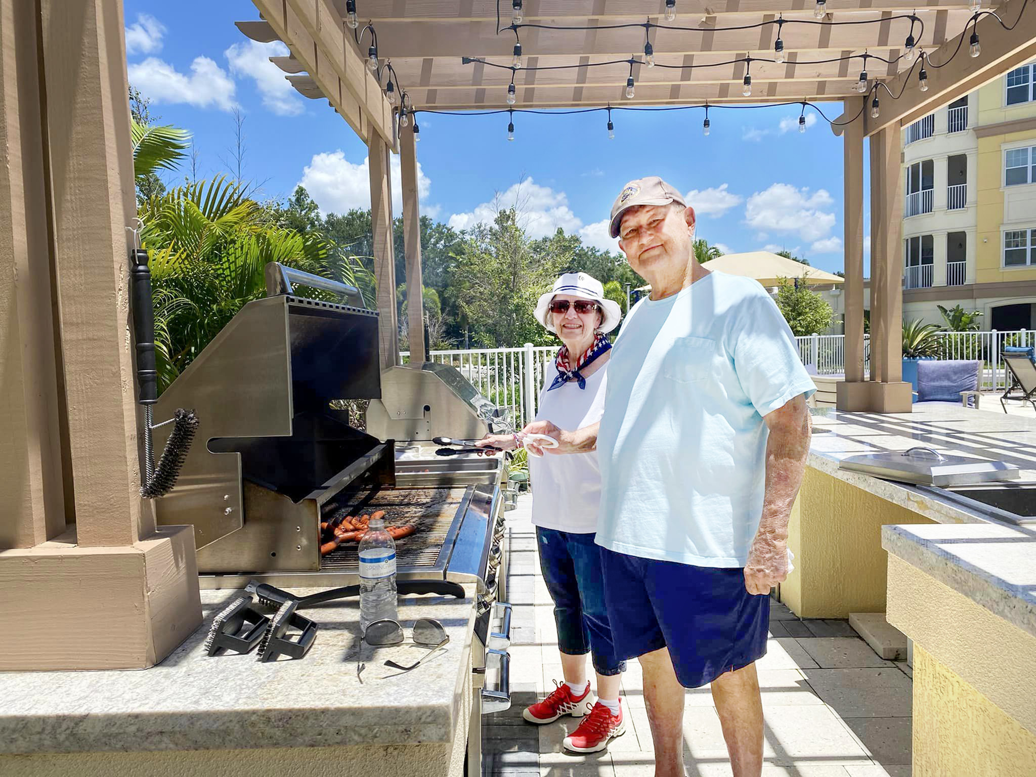 Two residents smiling at the camera while using the outdoor grill at the Alloro at University Groves on a bright and sunny day