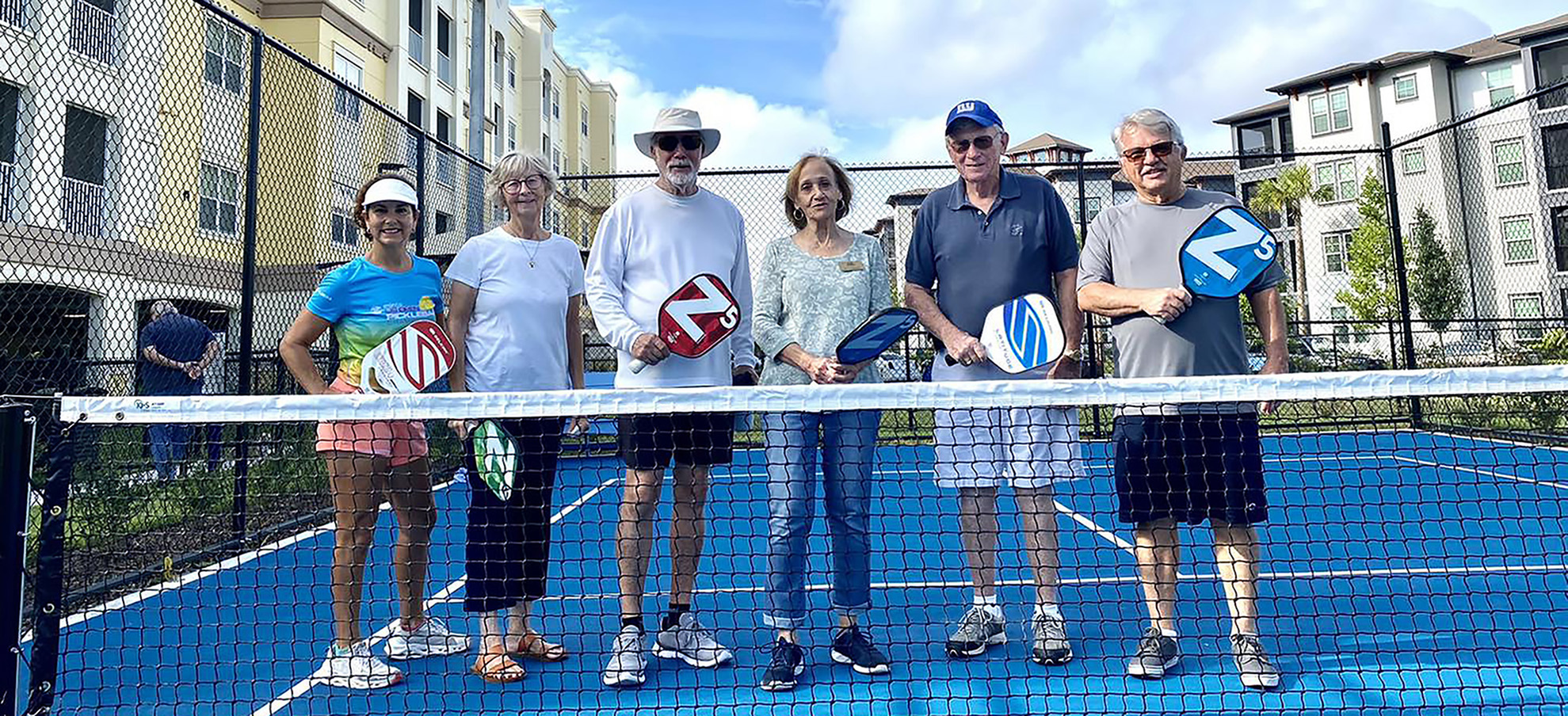 6 residents posing and smiling on the pickleball court while holding pickleball paddles