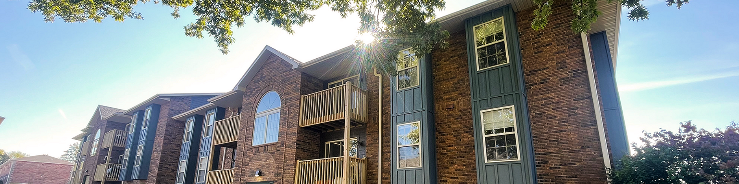 Charming apartment building with brick facade, surrounded by lush greenery and sunny blue skies, offering spacious balconies.