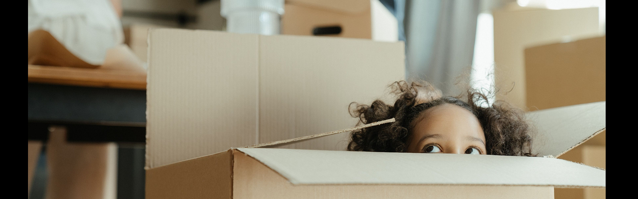 Charming scene of a child playfully peeking out of a moving box in a cozy, sunlit room, capturing the excitement of moving into a new home.