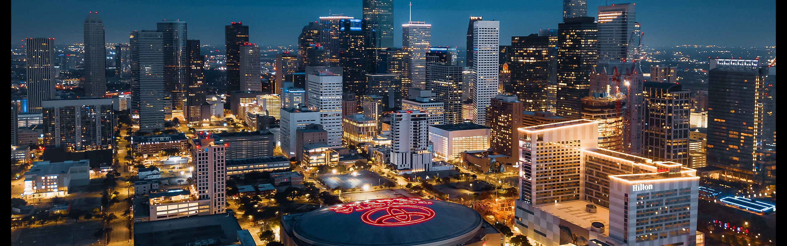 Vibrant aerial view of a bustling city skyline at dusk, with illuminated skyscrapers and a prominent arena glowing in the heart of downtown.